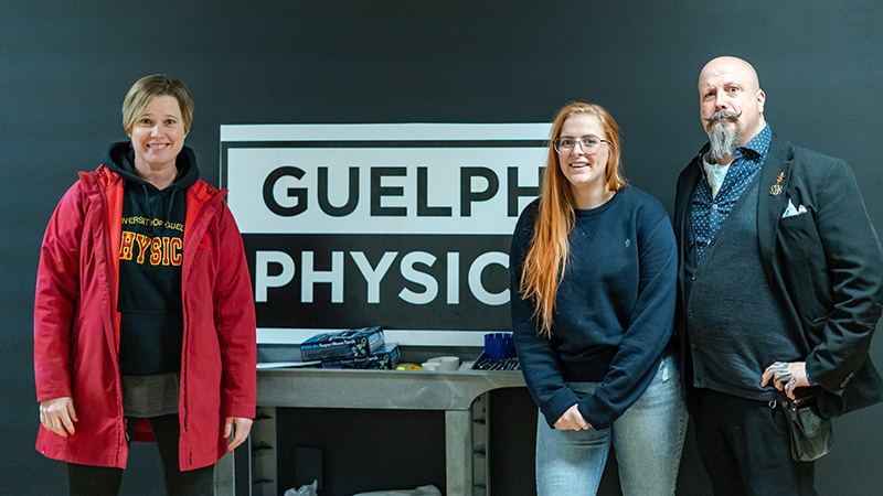 Joanne O'Meara, Samantha Buck and the Great Orbax posing in front of the Guelph Physics sign