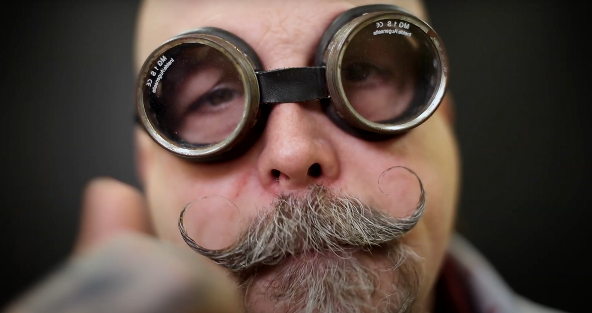 Close-up of a man with a curly moustache and goatee wearing goggles.