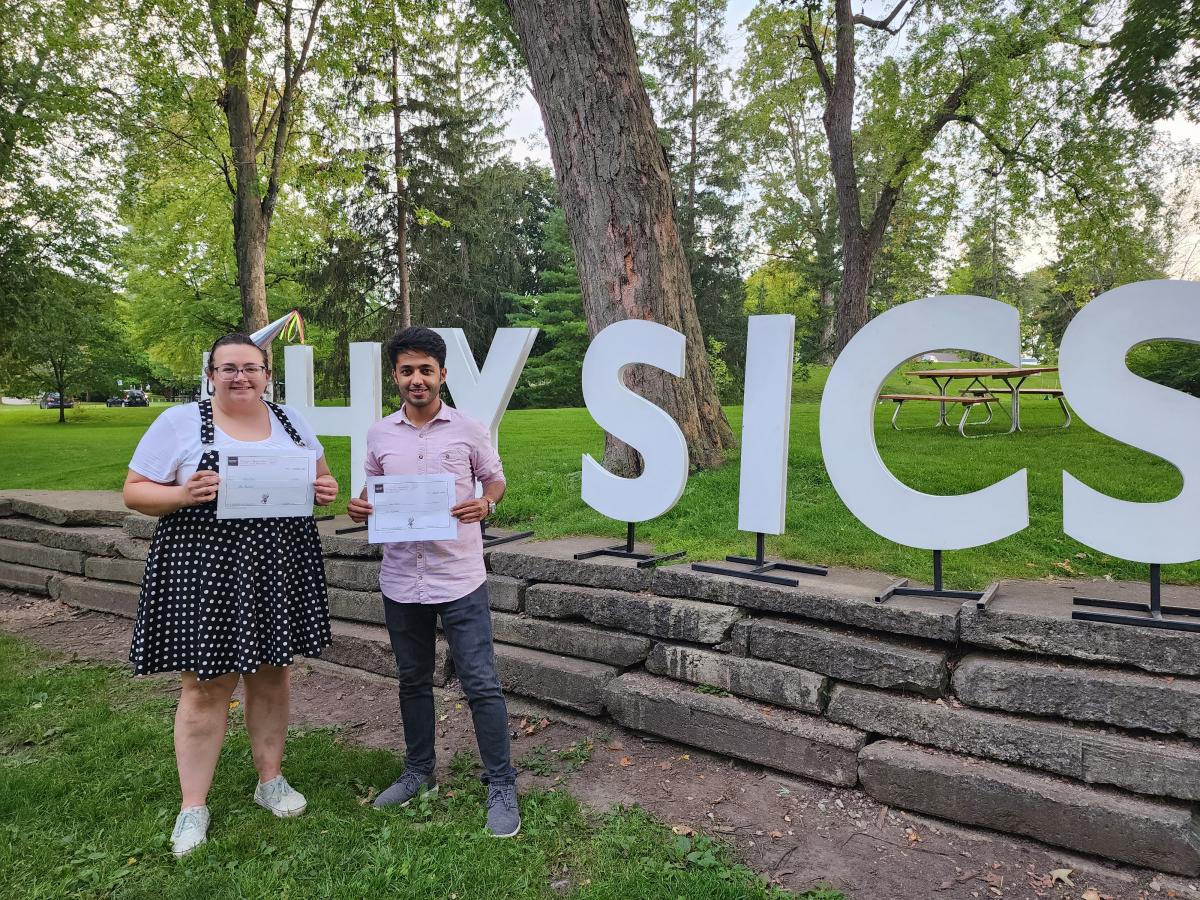 Two students standing in the park in front of large letters spelling Physics.