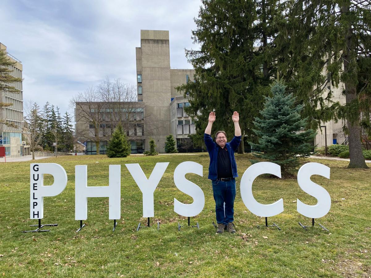 Man standing with raised arms in the I place, in oversized letters spelling Physics. 