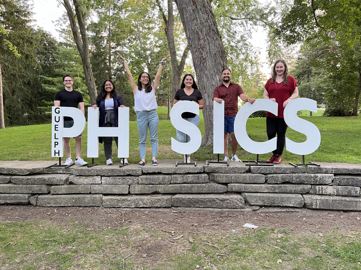 A group of graduate students are standing on a low stone wall in the park amongst letters spelling PHYSICS, one person is replacing the Y and standing with their arms up in a Y shape.