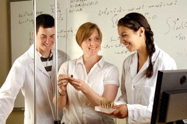Three people in a classroom with equations on the whiteboard behind them, doing an experiment with suspended weights